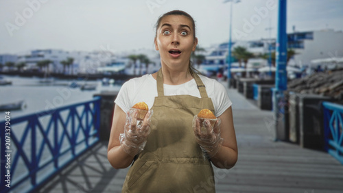 Bild auf Leinwand Woman holding two muffins with gloved hands in apron on a waterfront street pier with marina boats and railing; surprise offering