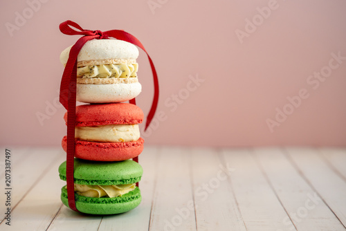 Colorful stack of three festive macarons tied with a red ribbon, creating a sweet dessert gift on a white wooden table with copy space against a pastel pink background