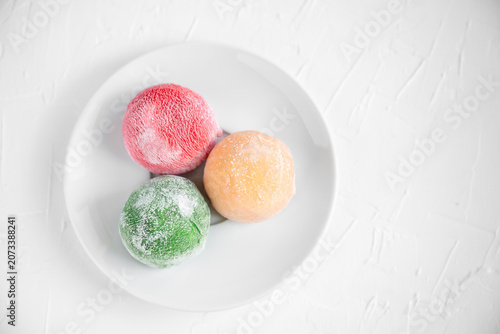 Three colorful mochi ice cream balls red, orange and green on a white plate over a textured white background, bright minimalist top view dessert for menus and food concepts