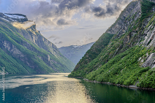 Geiranger, Majestic Norwegian Fjord Landscape, Dramatic fjord landscape with steep, green mountains and calm water under a cloudy sky. Serene natural beauty.