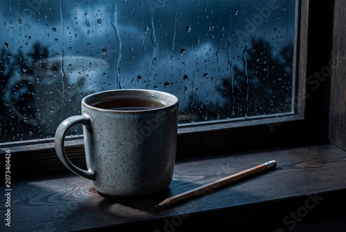 A premium commercial still life shot of a handcrafted ceramic mug filled with dark tea and a worn wooden pencil lying next to it on a dark open-pore wood windowsill during a storm.
