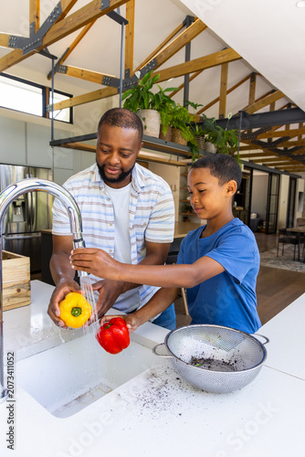 Billede på lærred African American father and son washing yellow, red bell peppers under metal fau