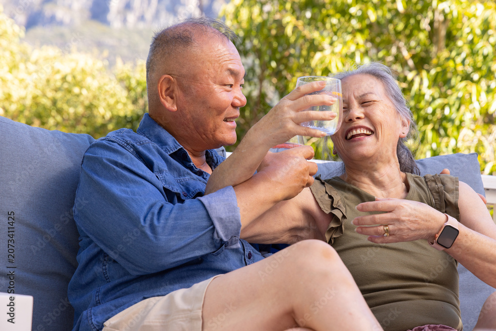 Fototapeta premium Senior Asian couple sitting on outdoor sofa, man holding clear glass, woman in olive top laughing