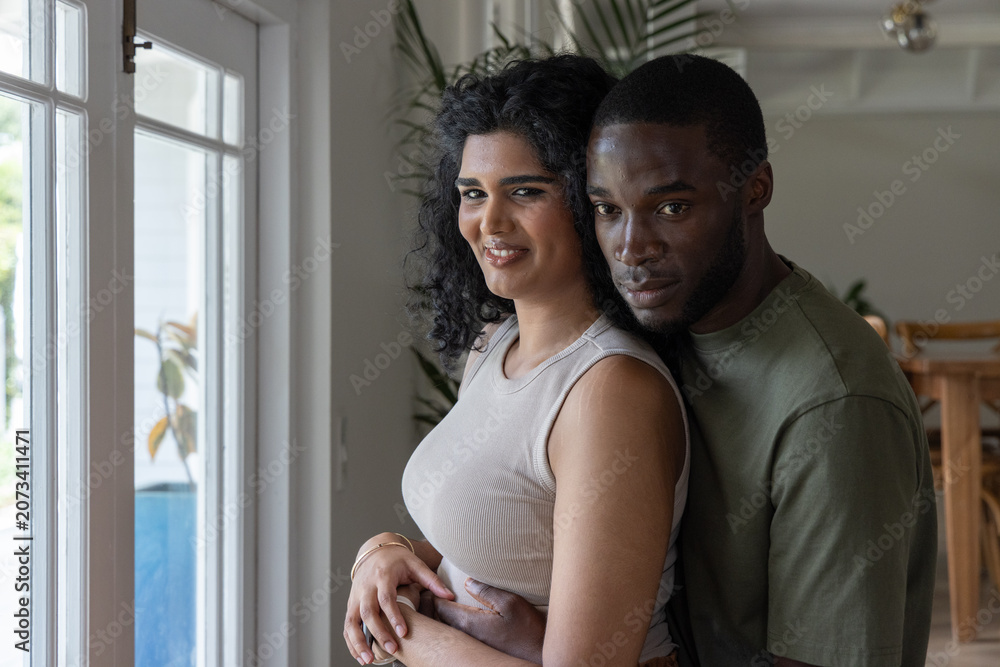 Fototapeta premium Diverse couple embracing by large glass door with potted plants, woman wearing beige sleeveless top