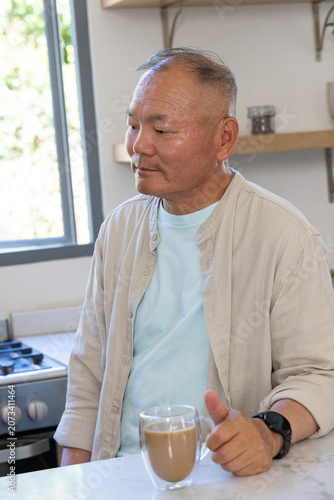 Senior Asian man holding double-walled glass mug and looking left at kitchen countertop window