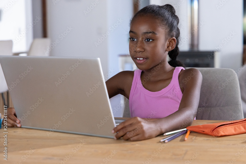 Obraz premium African American child in pink top sitting at dining table, using laptop with orange pencil case