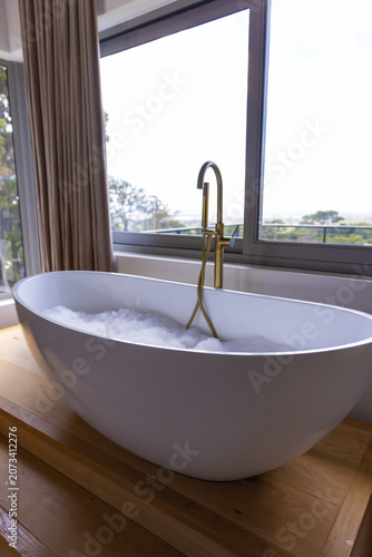 White freestanding tub is sitting on raised deck with bubbles beside brass faucet near large window