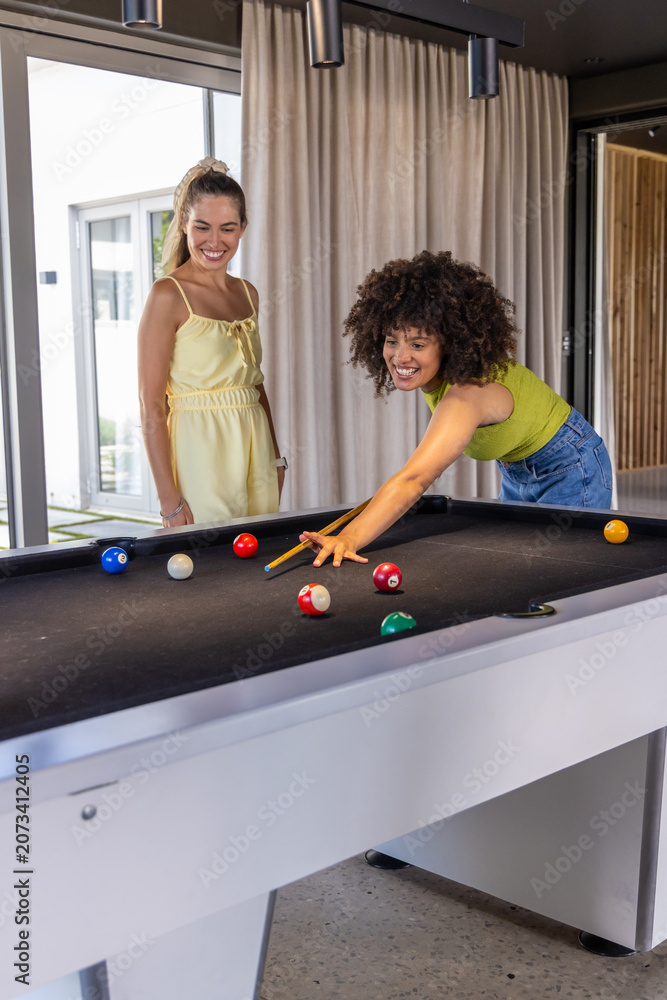 Fototapeta premium Diverse female friends playing pool in rec room, leaning over black-felt table using cue stick