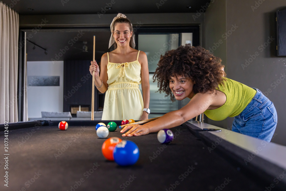 Naklejka premium Diverse female friends playing pool at home with smartphone and cues, lime top, yellow dress