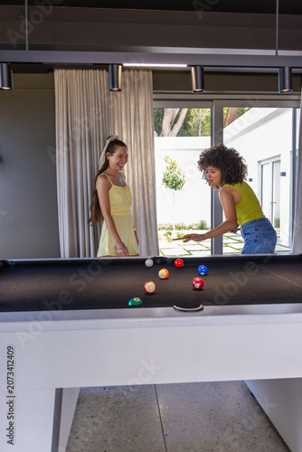 Diverse female friends playing pool in modern game room wearing yellow sundress and lime top, cue