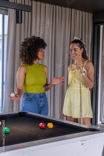 Two adult women standing chatting at modern lounge by black-felt pool table holding cue ball, cue
