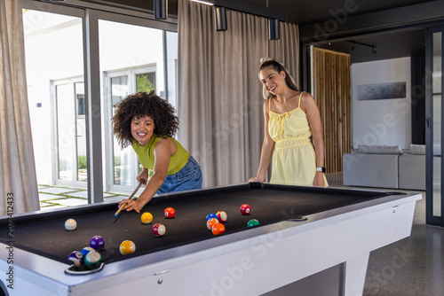 Diverse female friends playing pool at home lounge on white table with cue, balls, lime top