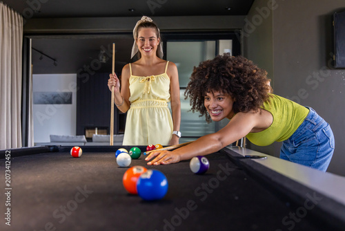 Diverse female friends playing pool at home with smartphone and cues, lime top, yellow dress