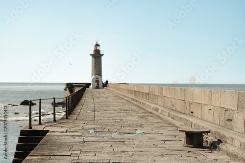 A solitary figure walks along a weathered pier, captivated by the view of a distant lighthouse
