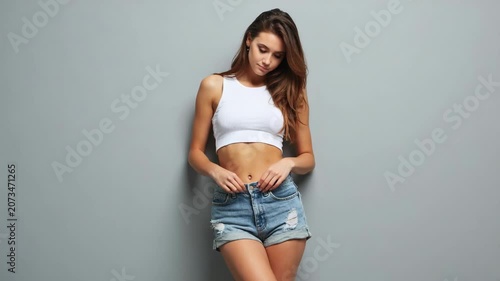 Confident young woman in white crop top and ripped denim shorts poses casually against gray background. Modern fashion, relaxed mood, studio lighting. checks how much weight she has lost or gained