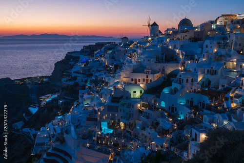 Fotografie Twilight cityscape of Oia’s whitewashed buildings and blue domes overlooking the Aegean Sea, with warm lights glowing at dusk