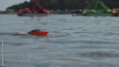 Slow motion video of small orange and black remote-controlled toy speedboat floating on the rippling blue water of a sea. The low-angle shot features a shallow depth of field, with colorful paddle