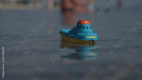 A small blue and yellow plastic toy boat floats on clear, calm water, casting a soft reflection. The low-angle shot captures the miniature vessel with a highly blurred background of distant watercraft