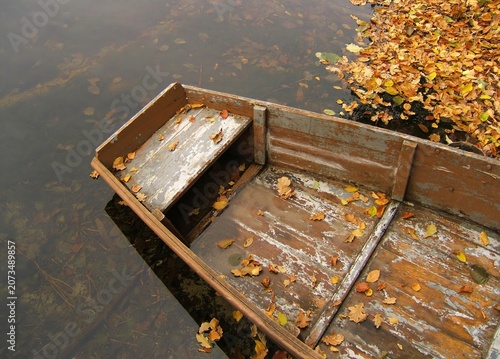 An old wooden rowing boat floating on a calm lake filled with fallen autumn leaves. The clear water reflects the surrounding trees and the peaceful atmosphere of the season.