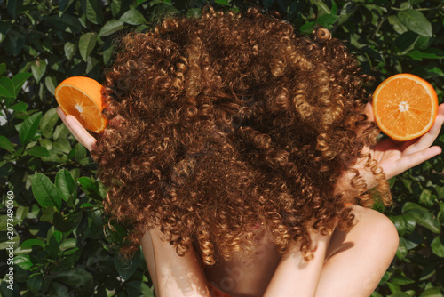 Foto Curly haired woman holds orange halves behind her head against leafy background