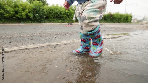 Close-up of a child wearing colorful rubber boots splashing through a rain puddle on a wet road. Water droplets fly through the air as small feet jump and play, capturing carefree childhood, outdoor