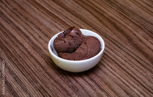 Two scoops of dark chocolate gelato in a white bowl on wood. Isolated rich dark chocolate ice cream in a minimalist white ceramic bowl. Dark rustic wooden surface with ample copy space for text.