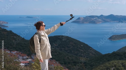 Female traveler taking a selfie video on a mountain top overlooking the sea. Beautiful woman hiker recording a video blog with an action camera on a selfie stick, standing with a view of the sea
