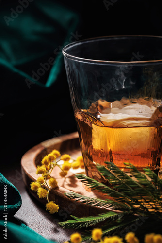 Close up whiskey or cognac in glass with ice on wooden tray decorated with mimosa flowers, elegant spring drink concept with dark background and copy space
