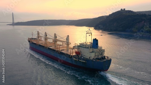 Photos Bulk carrier with deck cranes navigating strait toward suspension bridge at golden sunset with hilltop fortress in background