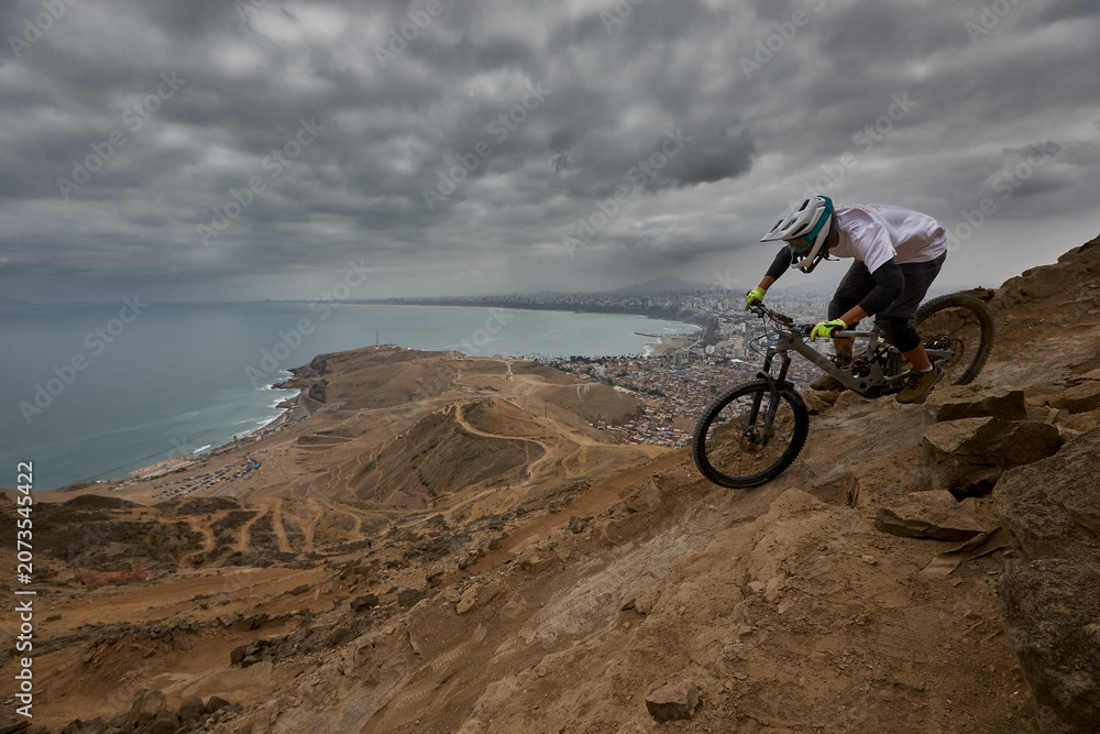 Fototapeta premium A seasoned athlete drops into a raw rocky descent at El Morro Solar, with the entire city of Lima and the Pacific Ocean laid out below, a backdrop that turns every run into something cinematic.