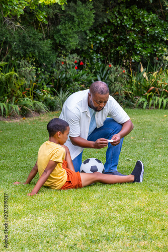 African American father and son kneeling, tending boy's leg in backyard with soccer ball, bandage