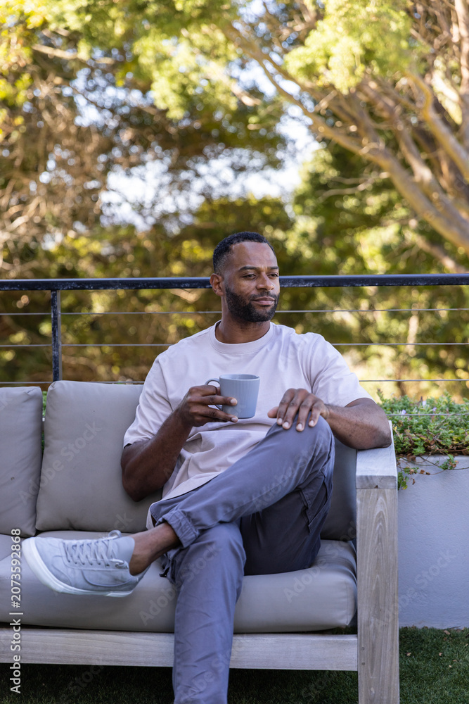 Obraz premium Mature African American man sitting on cushioned patio sofa holding ceramic mug, wearing T-shirt