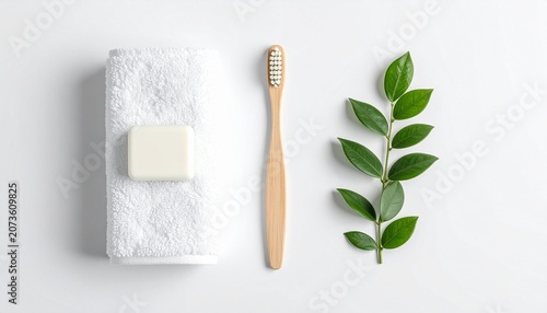 A neatly arranged bathroom setup featuring a folded white towel with a bar of soap on top, a wooden toothbrush, and a sprig of green leaves on a plain surface.