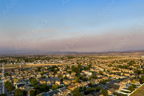 Aerial view of a suburban residential neighborhood with rows of houses, green trees, and local streets under a hazy sky caused by distant brushfire smoke, Lehi, Utah, United States.
