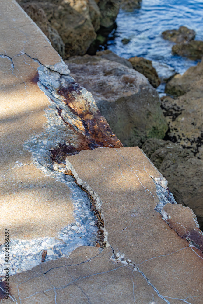 Fototapeta premium High angle view of severely cracked and spalling concrete seawall with rust stains and exposed aggregate next to rocky ocean shore