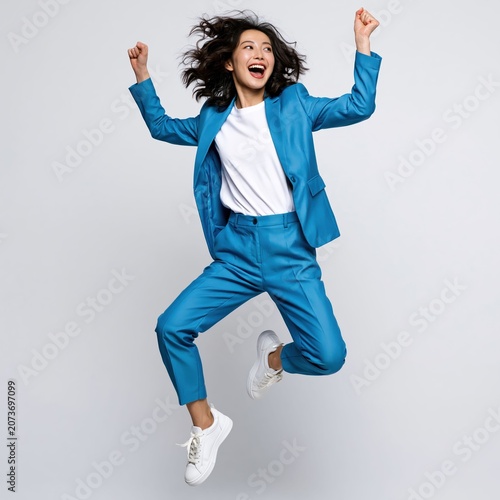 Asian woman jumps in front of a white background wearing a blue suit in a joyful and energetic pose.