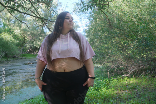 Plus size woman stands beside a quiet forest stream, wearing casual sportswear and gazing into the distance, expressing confidence, self acceptance and connection with nature