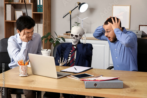 Schilderij op canvas Stressed male colleagues with human skeleton at table in office