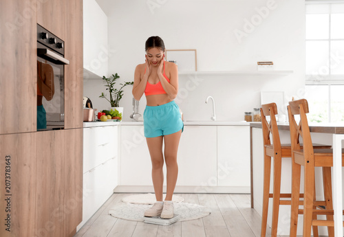 Happy young Asian woman measuring weight on scales in kitchen. Diet concept