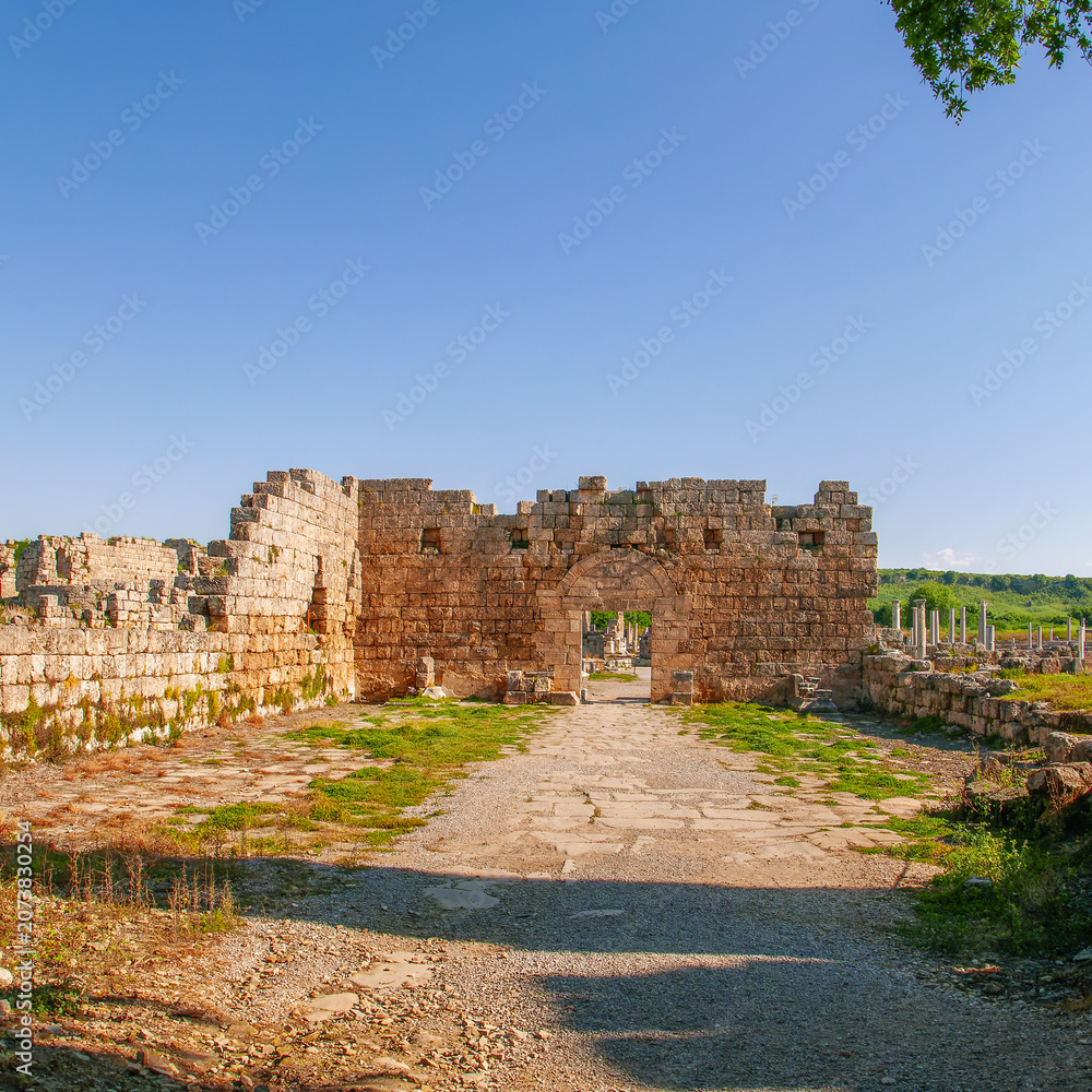 Fototapeta premium A scenic view of the ancient stone ruins and arched gateway at the historic site of Perge in Antalya, Turkey, under a clear blue sky.