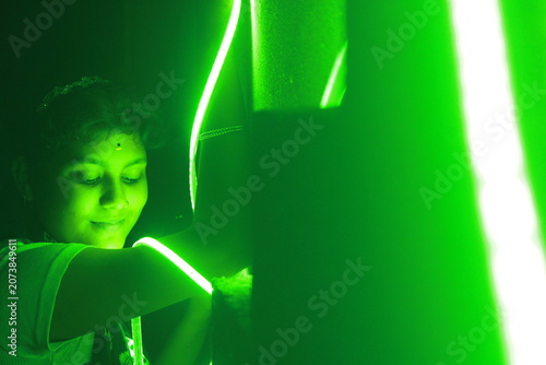 Creative portrait of a young Indian girl illuminated by vibrant green neon LED lights against a dark background. The dramatic lighting creates a futuristic atmosphere