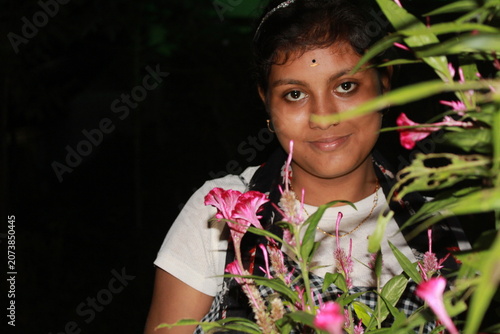 Portrait of a young Indian woman posing beside flowering plants in an outdoor garden at night. The image captures natural beauty, confidence, and contemporary Indian lifestyle, with vibrant flowers
