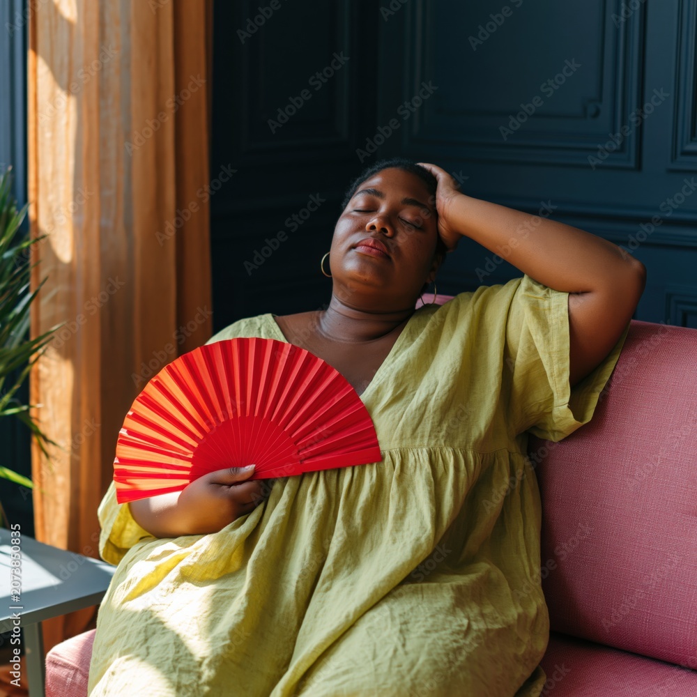 Fototapeta premium Relaxed woman fanning herself on pink sofa with red folding fan for summer lifestyle portrait and hot weather relaxation background