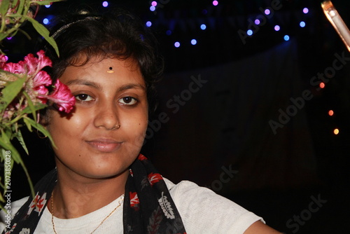 Portrait of a young Indian woman posing beside flowering plants in an outdoor garden at night. The image captures natural beauty, confidence, and contemporary Indian lifestyle, with vibrant flowers