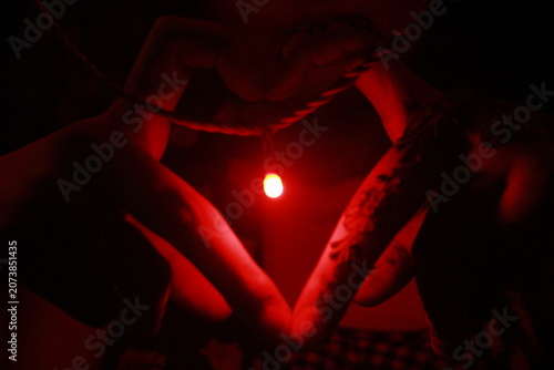 Creative low-light image of hands forming a heart shape around a glowing red LED light against a black background. The warm red illumination creates a dramatic and romantic atmosphere