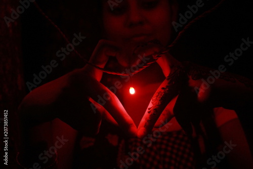Creative low-light image of hands forming a heart shape around a glowing red LED light against a black background. The warm red illumination creates a dramatic and romantic atmosphere