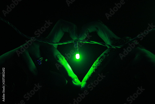 Creative close-up image of hands forming a heart shape around a glowing green LED light against a dark background. The composition symbolizes love, hope, connection, energy, and modern lighting.