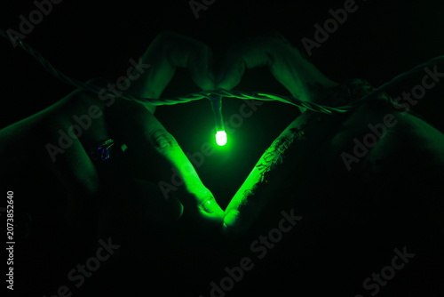 Creative close-up image of hands forming a heart shape around a glowing green LED light against a dark background. The composition symbolizes love, hope, connection, energy, and modern lighting.