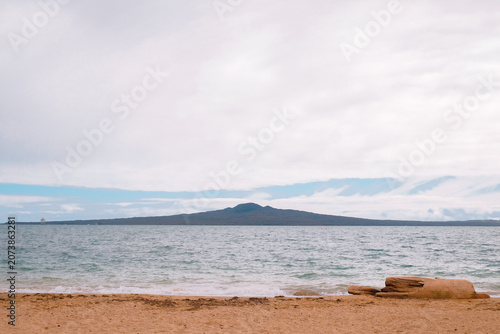 Auckland, New Zealand: scenic view of volcanic Rangitoto Island silhouette across ocean water from sandy beach shore under overcast cloudy sky.