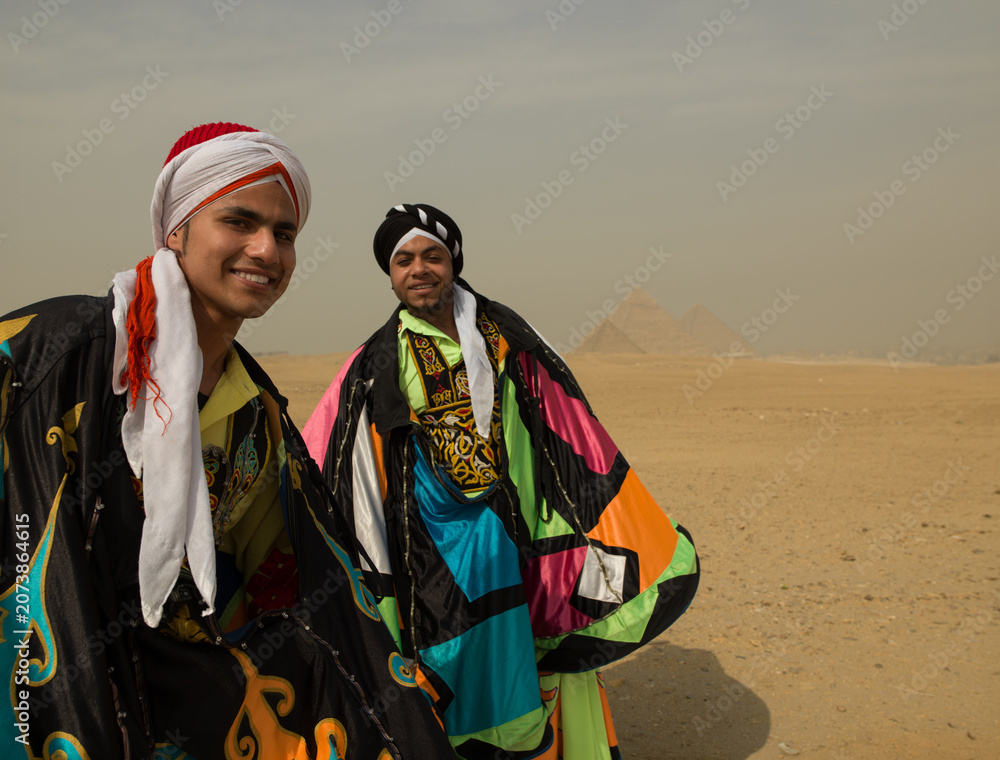 Fototapeta premium Two cheerful young Egyptian folk dancers in colorful tanoura costumes and turbans smiling before the Giza pyramids ideal for cultural festival and